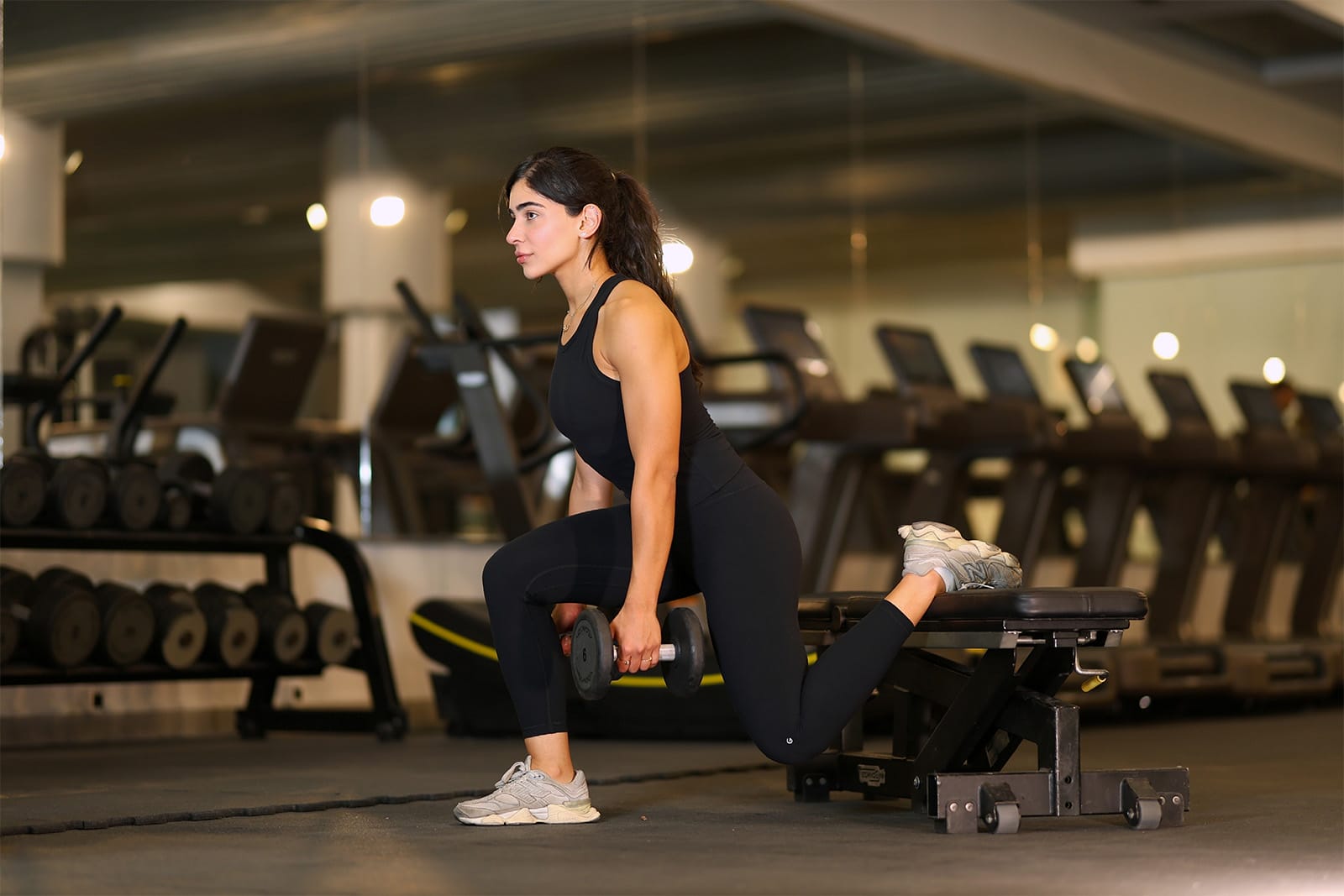 A woman in black athletic wear performs a Bulgarian split squat in a gym, holding dumbbells. Her back foot rests on a bench, and her front knee is bent at a right angle. Treadmills and free weights are visible in the background.