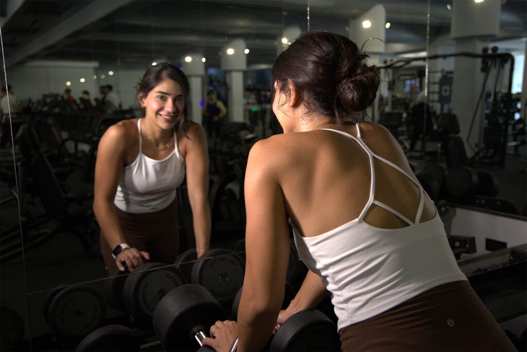 A woman in athletic wear leans on a rack of dumbbells in a gym, looking at her reflection in the mirror and smiling. The gym is spacious with various equipment visible in the background, and the lighting is bright.