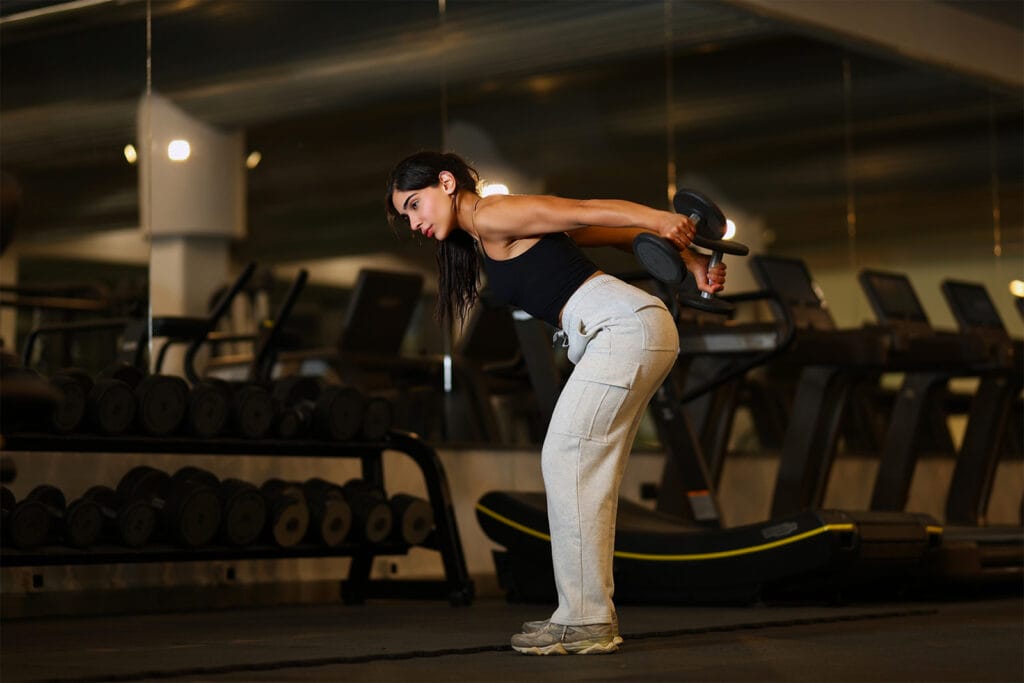 A woman in athletic wear stands in a gym, performing a bent-over triceps kickback with dumbbells. She faces sideways, knees slightly bent, arms extended back, with treadmills and a rack of weights in the background.