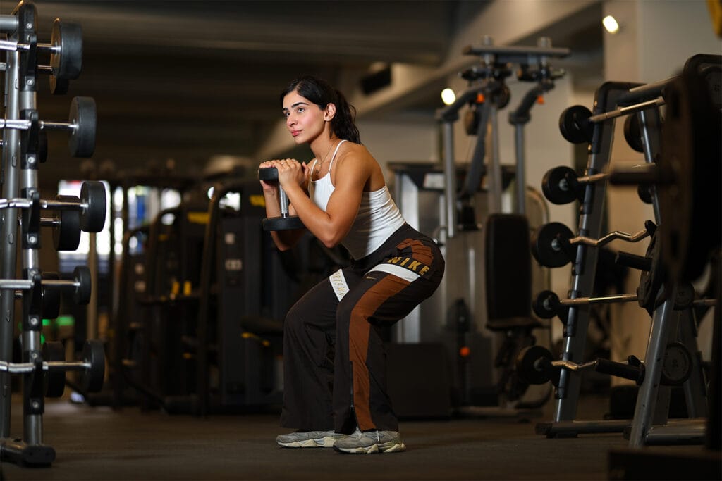 A woman in workout clothes is performing a squat with a dumbbell held close to her chest in a gym. She is surrounded by weightlifting equipment and machines, with focused expression, mid-exercise, in a well-lit indoor fitness center.