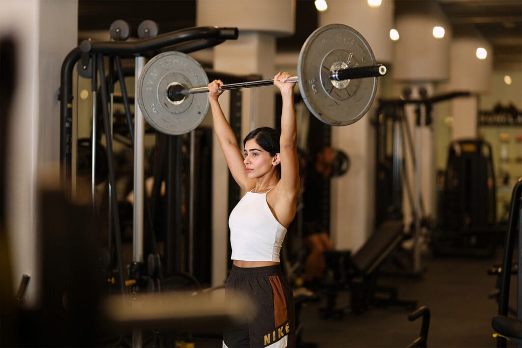 A woman in a white tank top and brown shorts lifts a barbell with weights overhead in a gym. She stands confidently with arms extended, surrounded by gym equipment and soft lighting in the background.
