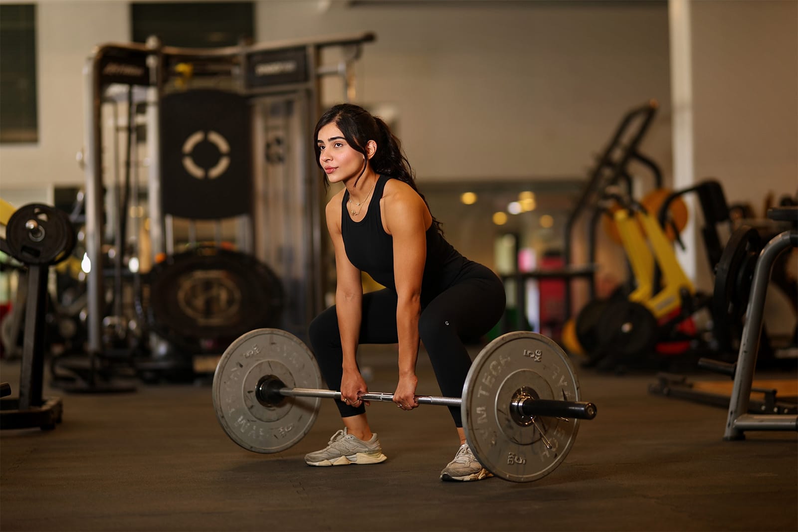 A woman in black athletic wear is preparing to lift a barbell in a gym. She is squatting with her hands gripping the bar, positioned for a deadlift. Exercise machines and weights are visible in the background, with gym lighting illuminating the scene.