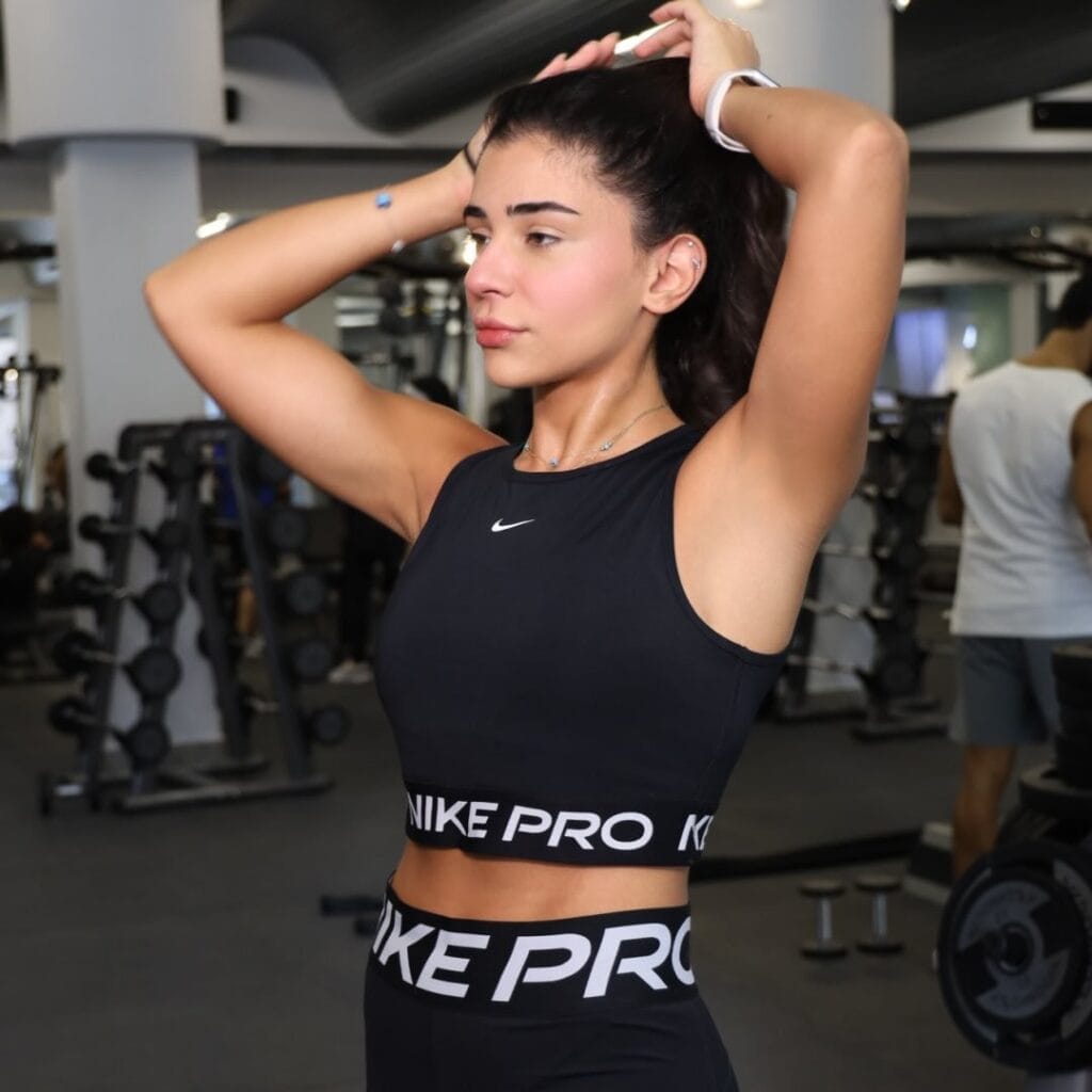 A woman in a black Nike Pro workout outfit stands in a gym, tying her dark hair back. Gym equipment and other people are visible in the background. She appears focused and ready to exercise.
